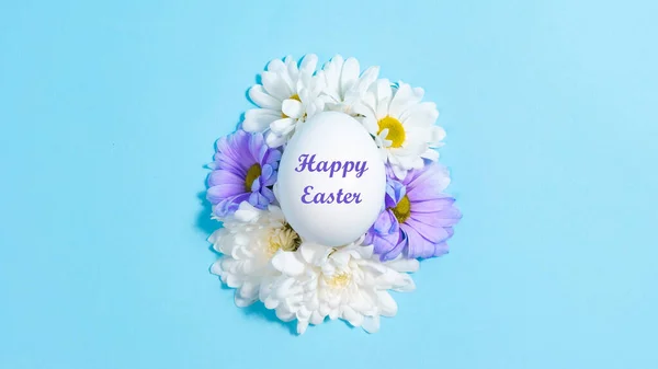 Stock image Happy Easter Holiday. Natural flowers of white and purple daisies near the white chicken egg. Blue background. Concept of natural food or Easter holiday. Greeting card.