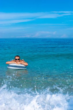 Bronz tenli, siyah saçlı bir adam, berrak mavi denizde sallanarak şişme yatakta dinleniyor. Deniz kenarında yaz tatili kavramı.
