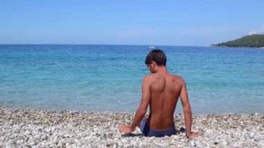 Siyah saçlı, gözlüklü genç adam bir çakıl taşı plajında oturur ve İyon Denizi 'nin berrak sakin sularına çakıl taşları atar. Yaz tatili deniz kenarı konsepti.