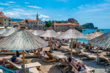 Budva, Karadağ - 4 Temmuz 2021: Eski şehir surları kalesinin arka planındaki Brijeg od Budva sahilindeki şemsiyeler altında dinlenen insanlar.