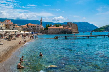 Budva, Karadağ - 4 Temmuz 2021: Eski kent surları kalesinin arka planındaki kumlu Brijeg od Budva plajında dinlenen insanlar. Eski Budva Kasabası kayalık bir yarımadada yer almaktadır. Yaz güneşli manzarası.