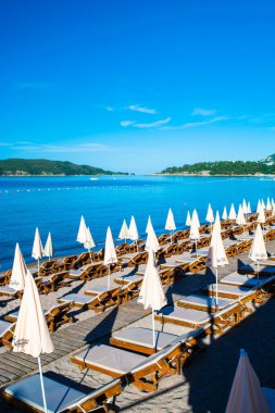 Becici, Karadağ - 02 Temmuz 2021: Güneşli yaz sabahlarında halk plajlarında güneş pansiyonları ve şemsiyeler. Skojl Adası geçmişi. Sakin deniz ve mavi gökyüzü. Budva Riviera. Deniz kenarı tatil köyü sezonu.