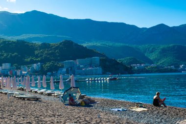 Becici, Karadağ - 02 Temmuz 2021: Yaz sabahı halk plajında güneş pansiyonları ve şemsiyeler. Arka planda Rafailovici binaları var. Sakin deniz. Mavi gökyüzü. Budva Riviera. Deniz kenarı tatil köyü sezonu.