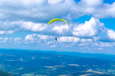 Eğitmenle paraşütle atlamak. Mavi bulutlu gökyüzü ve yeşil dağlardan oluşan güzel bir manzaranın arka planında bir paraglider silueti. Dağ manzaralı. Aşırı hobi ve macera.