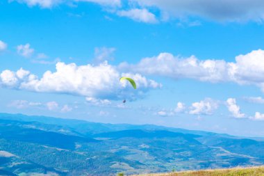Eğitmenle paraşütle atlamak. Mavi bulutlu gökyüzü ve yeşil dağlardan oluşan güzel bir manzaranın arka planında bir paraglider silueti. Dağ manzaralı. Aşırı hobi ve macera.