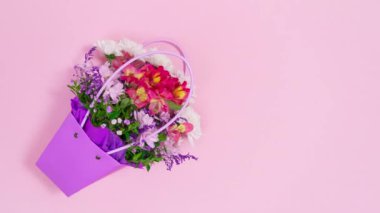 Pembe arka planda, beyaz kart ve hediye kutusuyla birlikte mor çantada bir buket taze çiçek görünüyor. Doğum günü kartı, anneler günü, kadınlar günü ya da başka bir gün için tebrik kartı. Hareket animasyonunu durdur. Uzayı kopyala Düz Yat.