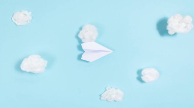 White paper airplane flies through white clouds on a blue background. Concept of travel, trips, air delivery. Flat lay. Copy space.