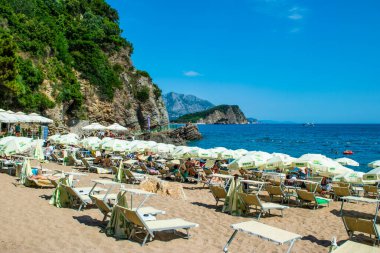 Morgen plajı, Karadağ - 4 Temmuz 2021: Budva yakınlarındaki kumlu plajda güneş pansiyonları ve şemsiyeler. Yaz güneşli manzarası.