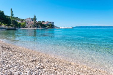 Serenely calm Adriatic Sea in summer season. Clean small-pebble beach early in the morning. Village of Promajna, Croatia. Houses at foot of foot on horizon. Vacation location. clipart