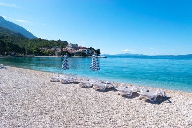 Yaz mevsiminde sakin Adriyatik Denizi. Çakıl taşlı sahilde güneş pansiyonları ve şemsiyeler. Hırvatistan 'ın Promajna köyü. Ufuktaki ayak basan evler. Tatil konumu.