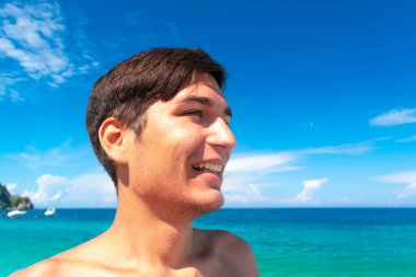 Young tanned smiling guy on the beach against a blue beautiful clean sea and sky. Concept of summer vacation by the sea, getting vitamin D while taking morning sunbaths. clipart