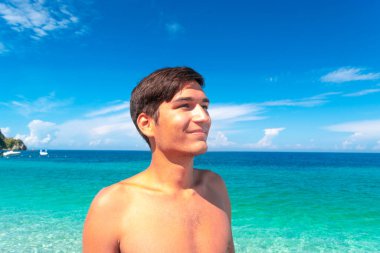 Young tanned smiling guy on the beach against a blue beautiful clean sea and sky. Concept of summer vacation by the sea, getting vitamin D while taking morning sunbaths. clipart