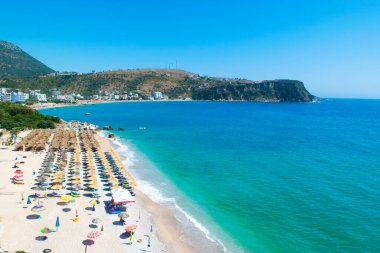 Himare, Arnavutluk - 15 Temmuz 2021: Güzel, temiz kum ve şemsiyeli çakıl taşı plajları ve güneş pansiyonları üzerindeki tatilciler. Güneşli dağlar mavi deniz manzarası. Himare. Arnavutluk. İyon Denizi. Üst Manzara.