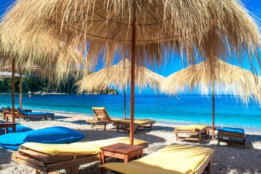 Saman plajı şemsiyeleri ve temiz kum ve çakıl taşı plajındaki rahat güneş şezlongları. Himare. Arnavutluk. İyon Denizi. Tatil deniz konsepti. Yaz güneşli deniz manzarası.