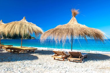 Saman plajı şemsiyeleri ve temiz kum ve çakıl taşı plajındaki rahat güneş şezlongları. Himare. Arnavutluk. İyon Denizi. Tatil deniz konsepti. Yaz güneşli deniz manzarası.