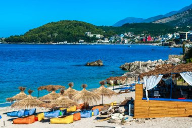 Güzel, temiz kum manzarası ve kumsal şemsiyeli çakıl taşı plajı ve dağ arkasındakiler. Himare. Arnavutluk. İyon Denizi. Tatil deniz konsepti. Yaz güneşli mavi deniz manzarası