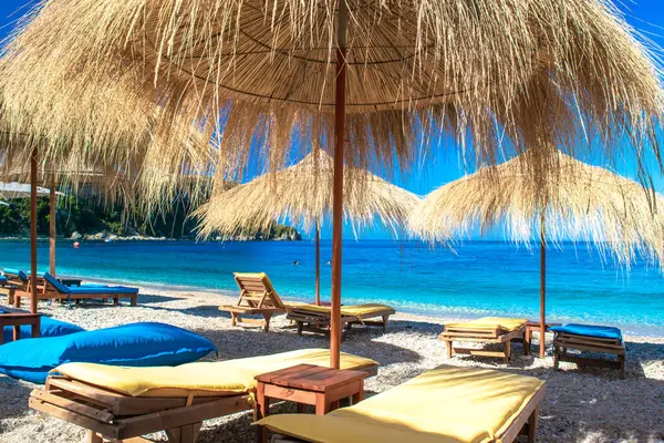 Saman plajı şemsiyeleri ve temiz kum ve çakıl taşı plajındaki rahat güneş şezlongları. Himare. Arnavutluk. İyon Denizi. Tatil deniz konsepti. Yaz güneşli deniz manzarası.