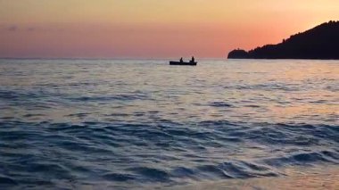 Calm sea surf in the foreground and a small boat with two fishermen against the pink sunset sky. Beautiful twilight seascape.