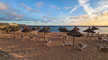 Gün batımında palmiye ağaçları, güneş ışıkları ve şemsiyeleri olan güzel, geniş kumlu bir sahil. Playa de las Americas, Tenerife, İspanya. Atlantik Okyanusu.