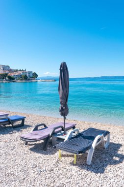 Yaz mevsiminde sakin Adriyatik Denizi. Çakıl taşlı sahilde güneş pansiyonları ve şemsiyeler. Hırvatistan 'ın Promajna köyü. Ufuktaki ayak basan evler. Tatil konumu.