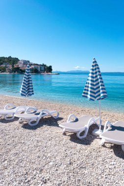 Yaz mevsiminde sakin Adriyatik Denizi. Çakıl taşlı sahilde güneş pansiyonları ve şemsiyeler. Hırvatistan 'ın Promajna köyü. Ufuktaki ayak basan evler. Tatil konumu.