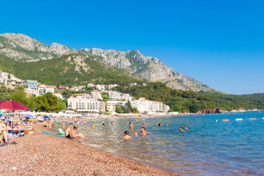 Sveti Stefan, Karadağ - 7 Temmuz 2021: Tatilciler Sveti Stefan adası yakınlarındaki plajda vakit geçiriyor. Güzel yaz güneşli yeşil dağ manzarası ve mavi gökyüzünde Adriyatik denizi.