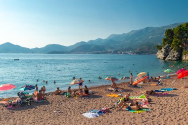 Sveti Stefan, Karadağ - 7 Temmuz 2021: Tatilciler Sveti Stefan adası yakınlarındaki plajda vakit geçiriyor. Güzel yaz güneşli yeşil dağ manzarası ve mavi gökyüzünde Adriyatik denizi.