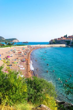Sveti Stefan, Karadağ - 7 Temmuz 2021: Tatilciler Sveti Stefan adası yakınlarındaki plajda vakit geçiriyor. Güzel yaz güneşli yeşil dağ manzarası ve mavi gökyüzünde Adriyatik denizi.