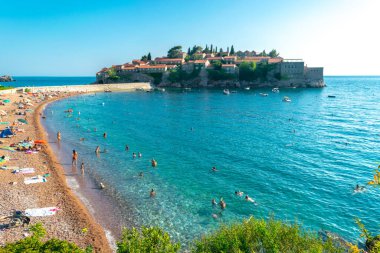 Sveti Stefan, Karadağ - 7 Temmuz 2021: Tatilciler Sveti Stefan adası yakınlarındaki plajda vakit geçiriyor. Güzel yaz güneşli yeşil dağ manzarası ve mavi gökyüzünde Adriyatik denizi.