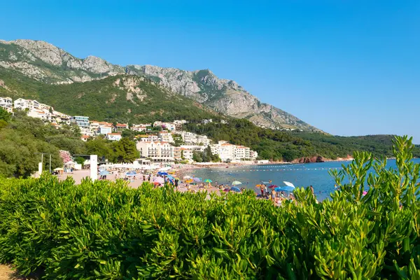 Sveti Stefan, Karadağ - 7 Temmuz 2021: Tatilciler Sveti Stefan adası yakınlarındaki plajda vakit geçiriyor. Güzel yaz güneşli yeşil dağ manzarası ve mavi gökyüzünde Adriyatik denizi.