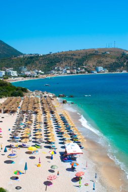 Himare, Arnavutluk - 15 Temmuz 2021: Güzel, temiz kum ve şemsiyeli çakıl taşı plajları ve güneş pansiyonları üzerindeki tatilciler. Güneşli dağlar mavi deniz manzarası. Himare. Arnavutluk. İyon Denizi. Üst Manzara.