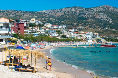 Himare, Arnavutluk - 15 Temmuz 2021: Şehir halk plajı. Güzel, temiz kumsalda ve çakıl taşlı plajda tatilciler. Arka planında yeşil tepe olan güneşli mavi deniz manzarası. İyon Denizi.