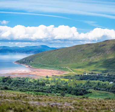 Kumlu körfezin manzarası, sakin deniz, mavi gökyüzü ve sisli mesafede Skye Adası, yeşil çimen kaplı tepeler, tepeler üzerinde kümülüs bulutları çizgisi.