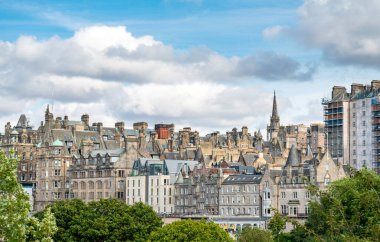 Edinburgh 'un güzel binaları ve çatıları, Princes Street Gardens' tan görünüyor, önplanda yeşil yaz ağaçları, mavi gökyüzü, tüylü beyaz bulutlar, yukarıdaki kilise kulesi.