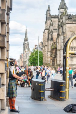 Edinburgh, İskoçya-30 Temmuz 2022: Geleneksel İskoç elbiseli bir İskoç boru sanatçısı, turistler ve geçenler için otobüsler, yaz aylarında, Sınır Bilim Festivali sırasında güvenlik bariyerlerinin yanında.
