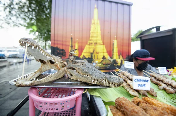 A Crocodile meat,street seller shows off two dried croc heads,mouths agape,with sharp teeth,next to various kebabs and other preparations of the meat,to attract customers.