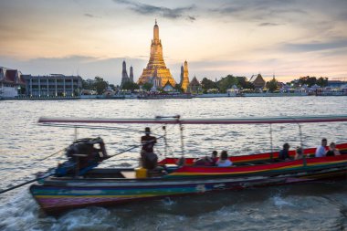 Alacakaranlıkta, bir Tayland Uzun Kuyruk teknesinin silueti tapınak kompleksinin altın pagoda 'larının yanından geçerken Bangkoks' un ana nehrinin zıt tarafından görülen açık günbatımı gökyüzünü işaret ederler..
