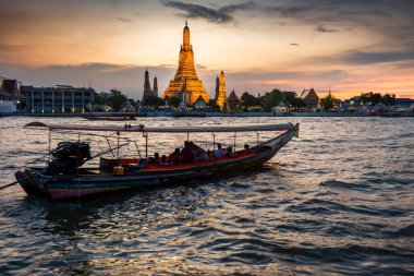 Alacakaranlıkta, bir Tayland Uzun Kuyruk teknesinin silueti tapınak kompleksinin altın pagoda 'larının yanından geçerken Bangkoks' un ana nehrinin zıt tarafından görülen açık günbatımı gökyüzünü işaret ederler..