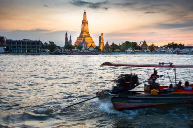 Alacakaranlıkta, bir Tayland Uzun Kuyruk teknesinin silueti kuruyor, tapınak şeklindeki görkemli füze kuleleri Bangkoks 'un karşı yakasından görünen açık günbatımı gökyüzünü gösteriyor..