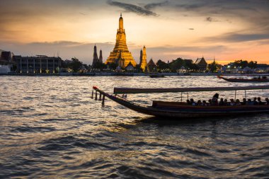 Alacakaranlıkta, bir Tayland Uzun Kuyruk teknesinin silueti tapınak kompleksinin altın pagoda 'larının yanından geçerken Bangkoks' un ana nehrinin zıt tarafından görülen açık günbatımı gökyüzünü işaret ederler..