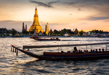 Alacakaranlıkta, bir Tayland Uzun Kuyruk teknesinin silueti tapınak kompleksinin altın pagoda 'larının yanından geçerken Bangkoks' un ana nehrinin zıt tarafından görülen açık günbatımı gökyüzünü işaret ederler..