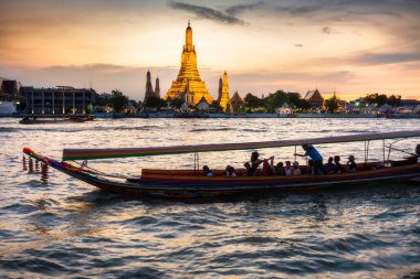 Alacakaranlıkta, bir Tayland Uzun Kuyruk teknesinin silueti tapınak kompleksinin altın pagoda 'larının yanından geçerken Bangkoks' un ana nehrinin zıt tarafından görülen açık günbatımı gökyüzünü işaret ederler..