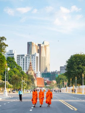 Phnom Penh, Kamboçya-23 Aralık 2023: Geleneksel turuncu cüppeler, keşişler, maske takan birçok kişi, güneşli bir öğleden sonra trafiğe kapalı, geniş bir yolda Kraliyet Sarayı Parkı 'nın önünden geçtiler..