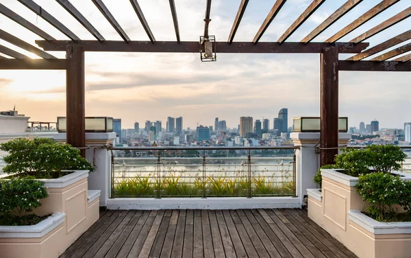 Framed by wooden beams of a high balcony,with decorative plants,the setting sun above distant high-rise buildings of Cambodia\'s capital city,reflecting sunlight from the surface of Tonle Sap River.