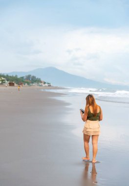 Akşam karanlığında Manila yakınlarında bir plajda, telefonunu selfie çekmek için tutarak, ılık sahil sularında amaçsızca geziniyor, pürüzsüz kumlar boyunca, eğleniyor, dans ediyor, hafif dalgaları tekmeliyor..