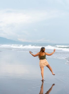 Akşam karanlığında Manila yakınlarında bir plajda, telefonunu selfie çekmek için tutarak, ılık sahil sularında amaçsızca geziniyor, pürüzsüz kumlar boyunca, eğleniyor, dans ediyor, hafif dalgaları tekmeliyor..