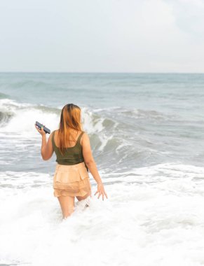 Akşam karanlığında Manila yakınlarında bir plajda, telefonunu selfie çekmek için tutarak, ılık sahil sularında amaçsızca geziniyor, pürüzsüz kumlar boyunca, eğleniyor, dans ediyor, hafif dalgaları tekmeliyor..