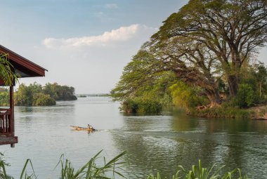 Nehir yatı nehrin yukarısına doğru gidiyor, günbatımına yakın, ön planda ayaklıkların üzerinde geleneksel Laos kulübesi, Mekong 'u sıcacık, yumuşak ışıkta saran güzel ağaç..