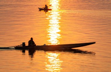 Mekong 'un sakin ve huzurlu sularında yüzen küçük teknelerde yaşayan insan figürlerinin siluetleri, batan güneşten, suya yansıyan altın ışık ışınları aracılığıyla..