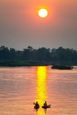 Kayaklardaki iki insan figürünün siluetleri Mekong 'un sakin ve huzurlu sularında yüzüyor. Güney Laos' taki güneşten gelen, suya, batan güneşe yansıyan altın ışık ışınları aracılığıyla..